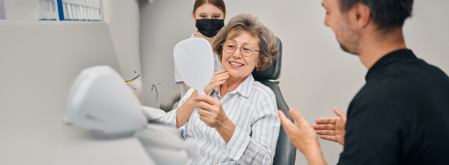 The image shows an elderly woman seated at a dental chair receiving dental care, with a dental professional standing nearby, both appearing to be engaged in a conversation.