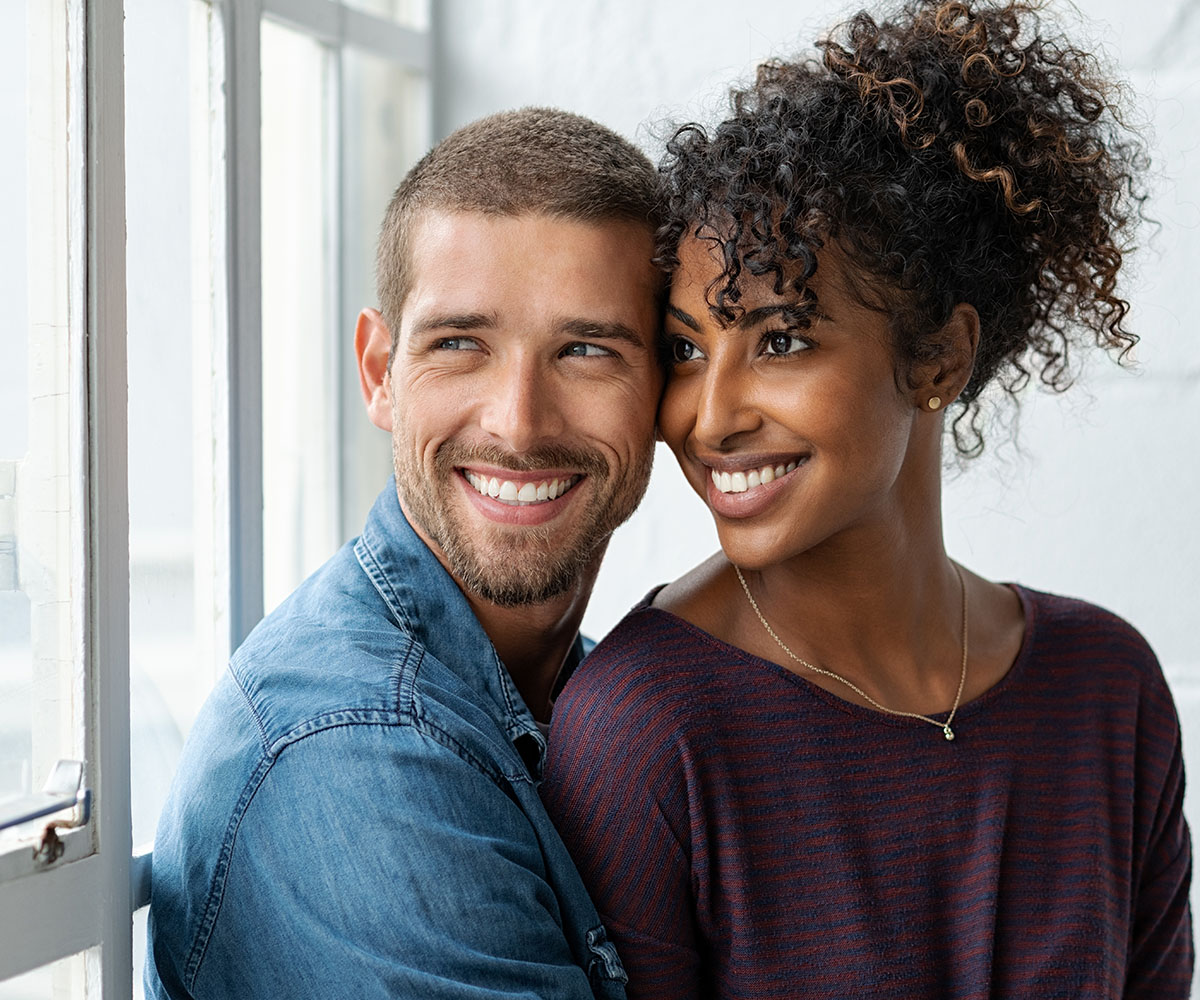 The image shows a man and a woman standing close together with smiles on their faces, looking directly at each other, against a window with daylight coming through.